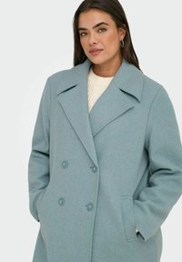 Gai zils vilnas mētelis ar dubulto pogu dizainu, plašu apkakli, sānu kabatām un teksturētu audumu. Pogas ir redzamas priekšpusē.