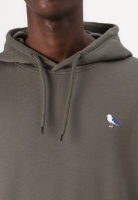Felpa verde oliva con cappuccio e coulisse, caratterizzata da un piccolo design ricamato di un uccellino bianco e blu sul davanti. Tessuto morbido.