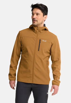 Man draagt een bruine jas met rits en capuchon en een zwarte broek, kijkt iets naar links tegen een effen achtergrond.