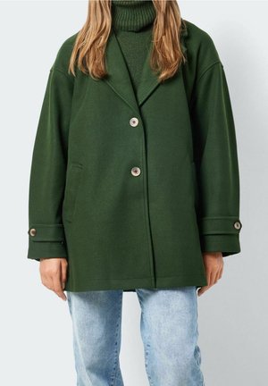 Kvinde iført mørkegrøn knaplukket frakke over matchende rullekravesweater og lyseblå jeans, stående mod en ensfarvet lys baggrund.