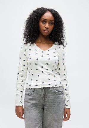 Γυναίκα με σγουρά μαλλιά που φοράει λευκό πουλόβερ με μακριά μανίκια και μικρά λουλούδια, μαζί με γκρι τζιν, όρθια μπροστά σε απλό φόντο.