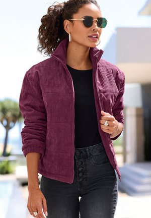 Woman wearing round sunglasses, hoop earrings, a purple corduroy jacket, black top, and high-waisted black jeans outdoors in daylight.