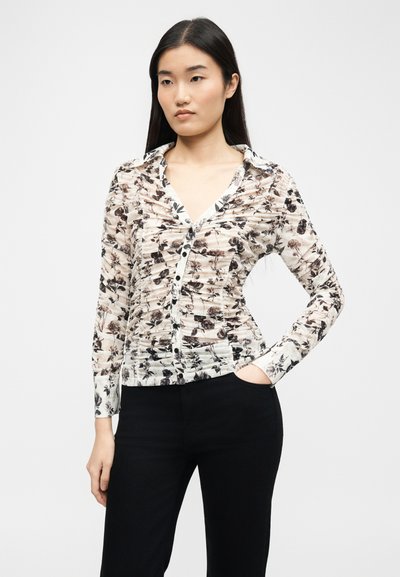 Γυναίκα φορώντας ένα ρουσέ λουλουδάτο πουκάμισο με κουμπιά και μακριά μανίκια, και μαύρο παντελόνι, στέκεται με το ένα χέρι στην τσέπη.