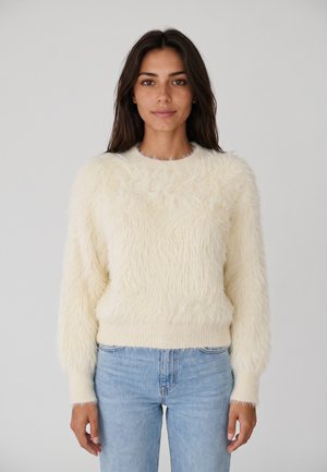 Donna con capelli lunghi e scuri che indossa un maglione morbido color crema e jeans azzurri chiari su uno sfondo grigio chiaro uniforme.