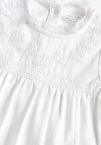 Vit babyklänning med broderad blommönster, spetsdetaljer vid halsringning och livstycke, samt samlad tyg för en mjuk och slät känsla.