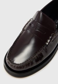 Braune Leder-Loafers mit glänzendem Finish, die seitlich geflochtene Details und eine kontrastierende schwarze Gummisohle aufweisen.