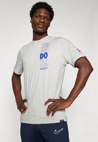 Mies seisoo kädet lanteilla päällä harmaa "JUST DO IT." -t-paita ja tummansiniset Nike-sortsit vaaleaa taustaa vasten.
