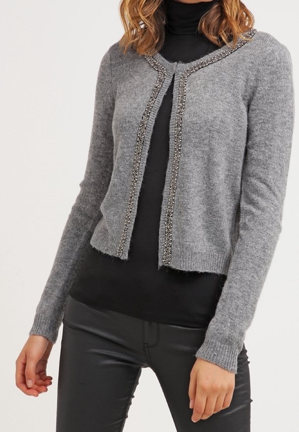Cardigan gris en matière douce, avec une coupe courte, des accents perlés sur le devant et des manches longues. Porté par-dessus un haut noir.