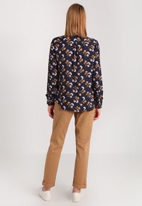 Granatowa koszula z długim rękawem w kolorowy kwiatowy wzór, z guzikami przy mankietach i plisą z tyłu, zestawiona z jasnobeżowymi spodniami z podwiniętymi nogawkami i białymi butami.