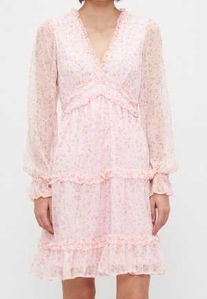 Femme portant une robe florale rose clair avec des manches longues transparentes, une bordure à volants et un décolleté en V, se tenant devant un fond uni.