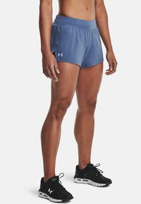 Idrottare som bär blå Under Armour träningsshorts och svarta Under Armour löparskor, ståendes på en vit bakgrund.