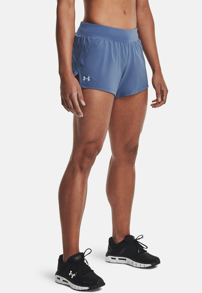Idrottare som bär blå Under Armour träningsshorts och svarta Under Armour löparskor, ståendes på en vit bakgrund.