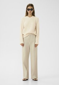 Un pull v-neck couleur crème associé à un pantalon large beige. Porté avec des lunettes de soleil noires et des sandales noires simples. Textures lisses tout au long.