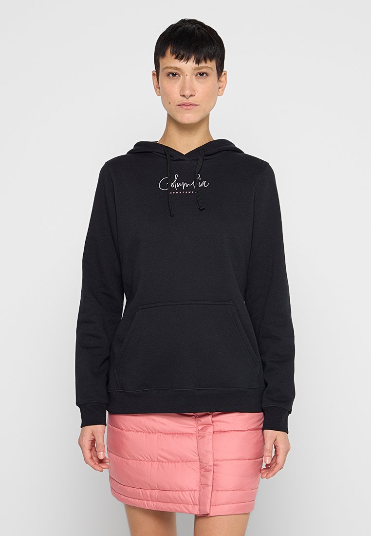 Schwarzer Kapuzenpullover mit einer Fronttasche, der ein weißes Logo aufweist. Glatte Textur, schlichtes Design. Kombiniert mit einem pinken, puffigen Rock.