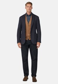 Mornarska blazerica iz teksturiranega materiala, rjav brezrokavnik in jeansa s kratkimi rokavi; v kombinaciji s temnimi kavbojkami in rjavimi semiš čevlji, kar ustvari slojevit videz.