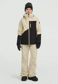 Skiausrüstung mit einer beige-schwarzen wasserdichten Jacke mit Reißverschlusstaschen und passenden Hosen, ergänzt durch schwarze Handschuhe und Skibrille.