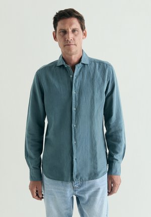 Hombre que lleva una camisa de manga larga color verde azulado con botones y vaqueros azul claro, de pie delante de un fondo claro y liso.
