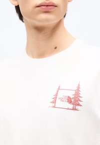 Κοντινό πλάνο ενός ατόμου που φοράει λευκό μπλουζάκι με ροζ γραφικό δύο δέντρων και το κείμενο "THE NORTH FACE" στο στήθος.