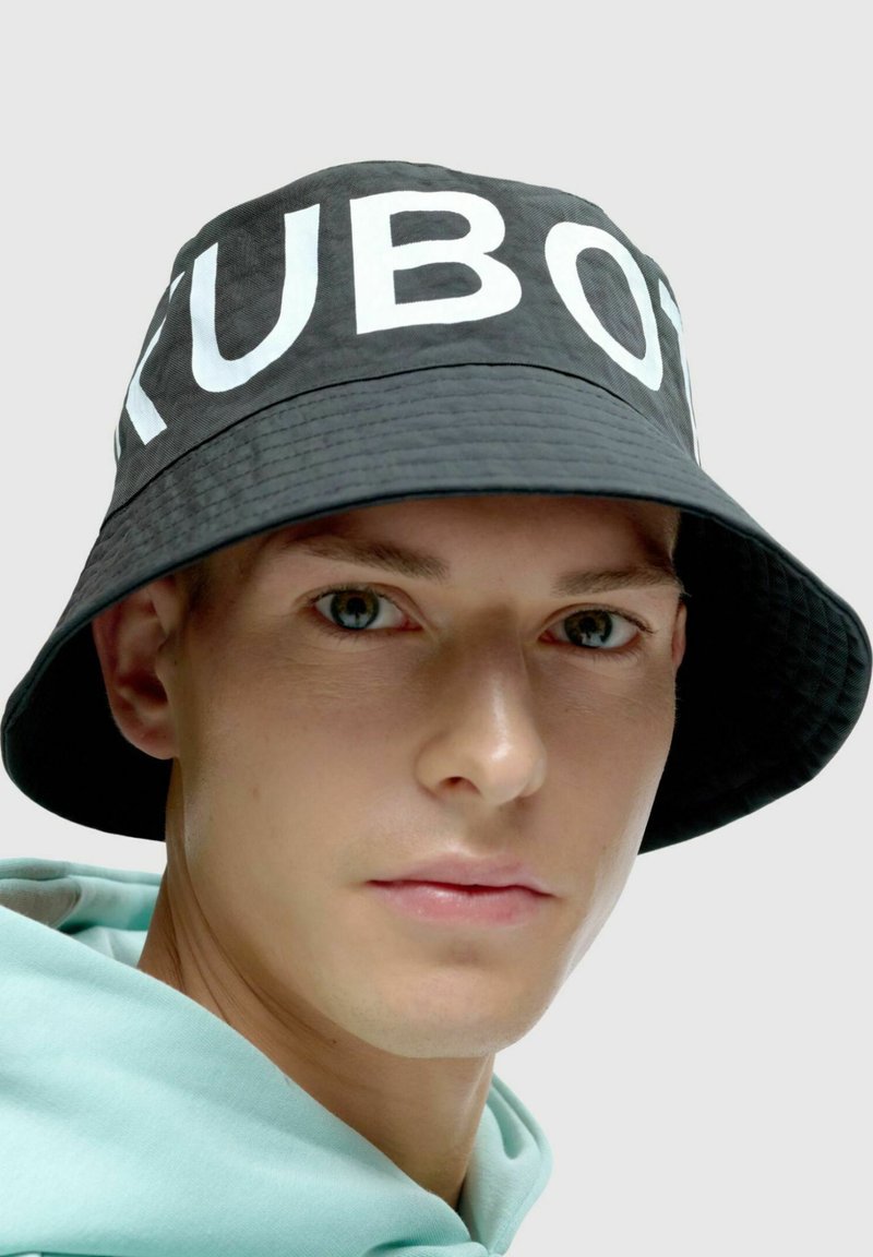 Czarny kapelusz typu bucket z białym napisem "KUBOT". Miękka tkanina, szeroki rondo i strukturalny design. Noszony przez osobę w jasnoniebieskiej bluzie z kapturem.