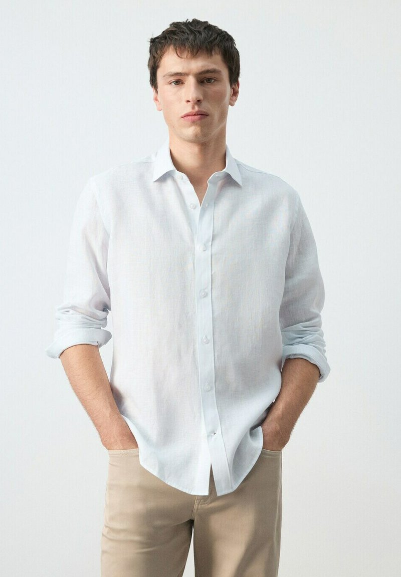 Jeune homme aux cheveux courts et foncés, portant une chemise blanche à boutons aux manches retroussées et un pantalon beige, les mains dans les poches, fond neutre.