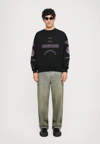 Hanorac negru cu text grafic purpuriu. Blugi verzi deschis, cu croi lejer. Modelul poartă adidași negri și ochelari de soare. Design minimalist.