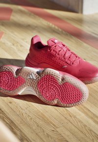 Red athletic shoes with textured mesh uppers and a cushioned sole featuring a wavy pattern. Outsole is light pink with nodules for grip.