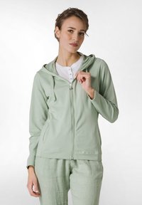 Sudadera con cremallera de color verde claro, hecha de algodón suave, que presenta una capucha, bolsillos frontales y una camisa blanca parcialmente abotonada debajo.