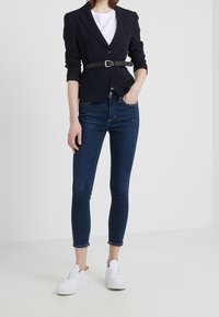 Blazer navy con un diseño ajustado, botón único y cinturón negro; combinado con jeans skinny azul oscuro y zapatillas blancas.