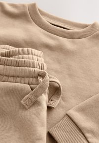 Hanorac bej din bumbac cu guler reiat, asortat cu pantaloni de aceeași culoare cu manșete elastice și talie cu șnur. Textură moale pe întreaga suprafață.