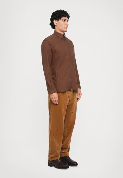Homme debout de profil portant une chemise brune zippée, un pantalon en velours côtelé léger marron clair ample avec de petits détails brodés, et des bottes brunes foncées.