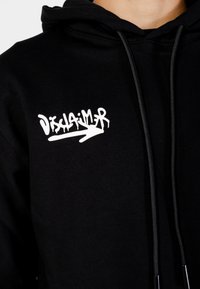Felpa nera con cappuccio, decorata con testo in stile graffiti bianco "Disclaimer" e una freccia sul petto, dotata di lacci neri per il cappuccio.