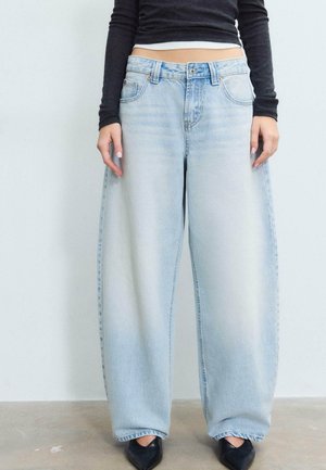 Oseba, ki nosi svetlo modre široke kavbojke, črno oprijeto kratko majico z dolgimi rokavi in črne koničaste čevlje, stoji na sivem tleh.