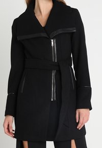 Cappotto in misto lana nero con ampio colletto caratterizzato da rifiniture in pelle, chiusura a zip e vita con cintura. Ha maniche lunghe e dettagli di design minimalisti.