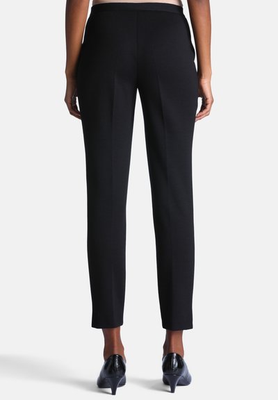Personne portant un pantalon noir sur mesure longueur cheville et des chaussures noires à talons, debout devant un fond clair uni.
