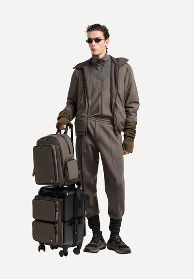 Completo sportivo marrone con giacca con zip, pantaloni coordinati e polsini in maglia testurizzata. Due borse sovrapposte in marrone e nero. Sneakers incluse.