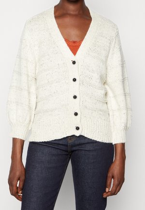 Cardigan blanc en maille épaisse avec boutons noirs porté sur un top en dentelle orange et un jean bleu foncé sur une personne debout.