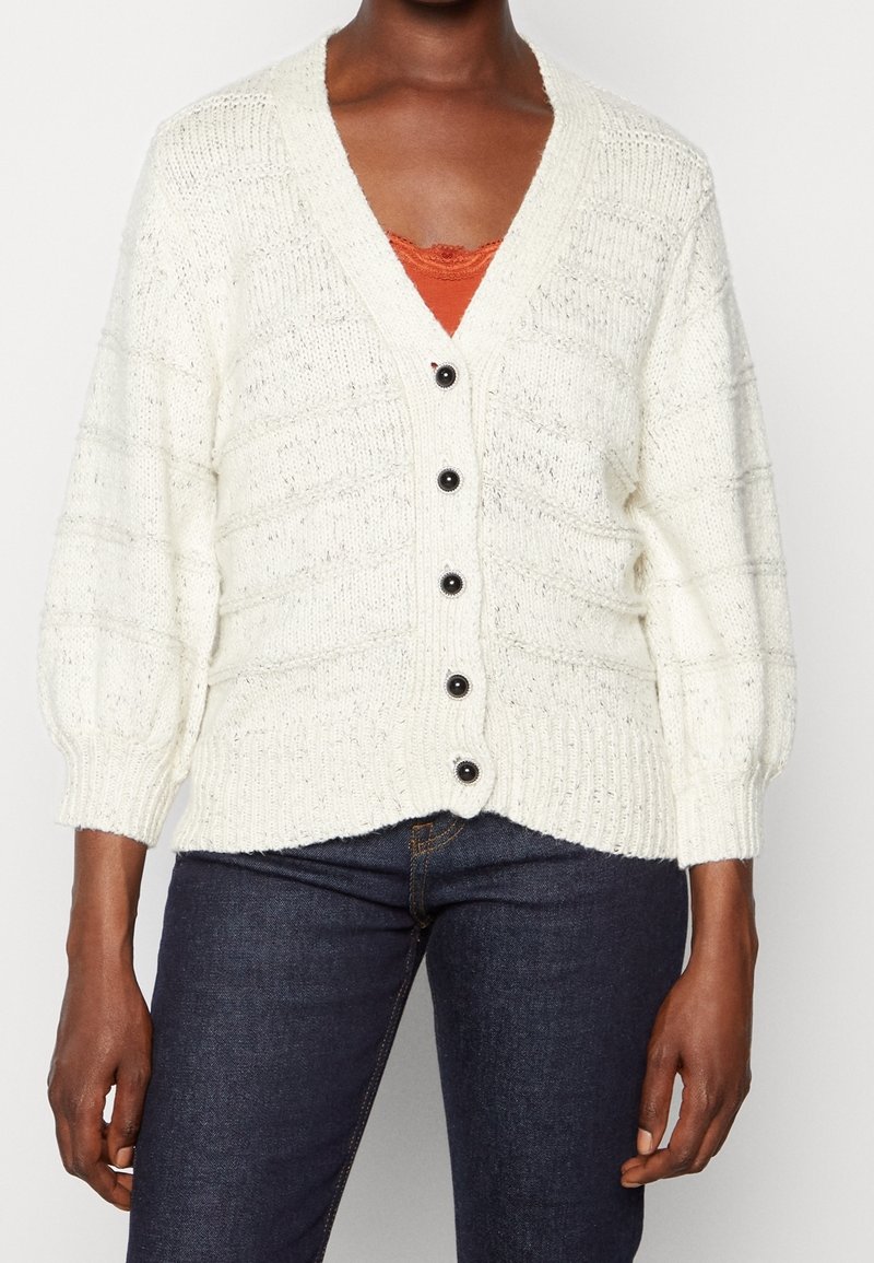 Cardigan blanc en maille épaisse avec boutons noirs porté sur un top en dentelle orange et un jean bleu foncé sur une personne debout.