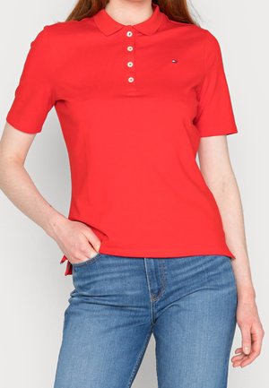 Personne portant un polo rouge à manches courtes avec des boutons et un jean bleu, debout avec une main dans la poche devant un fond uni.