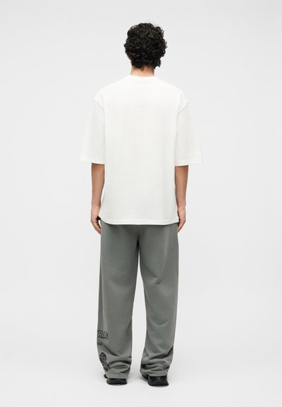 Άνδρας με σγουρά σκούρα μαλλιά στέκεται με την πλάτη στραμμένη φορώντας ένα άσπρο φαρδύ Τ-shirt, γκρι παντελόνι με φαρδιά μπατζάκια και μαύρα παπούτσια σε λευκό φόντο.