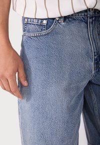 Lichtblauwe denim jeans met een gestructureerde afwerking, voorzien van een vijf-pocket ontwerp en metalen accessoires aan de taille, met een gedeeltelijk zichtbare hand.