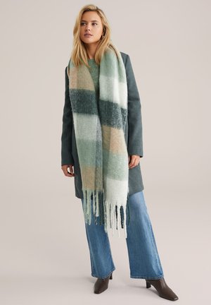 Sciarpa allungata in verde, crema e pesca con motivo a quadri, realizzata in materiale morbido con bordi frangiati; indossata sopra un cappotto grigio e jeans.