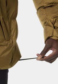 Olivově zelená péřová bunda s hladkým, lesklým povrchem, na které je ruka upravující elastický stahovací šňůru na spodním lemu, což ukazuje na praktičnost.