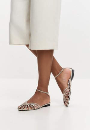 Barfodet person iført elegante flade sandaler med rhinstenremme og ankelspænde, kombineret med off-white bukser med brede ben.