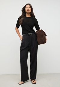 Top de punto negro con textura y mangas cortas, pantalones negros de cintura alta y bolso de ante marrón. La modelo lleva chanclas negras y está de pie frente a una pared clara.