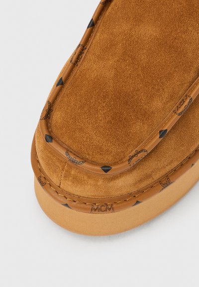 Chaussure à enfiler en daim marron avec une texture lisse, dotée d'une bordure en cuir avec logo et de subtils accents triangulaires noirs le long du bord.