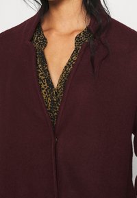 Bordeauxrode fleecejas met een rits aan de voorkant, met daaronder een blouse met luipaardprint. Opvallende texturen zijn zachte fleece en gladde stof.
