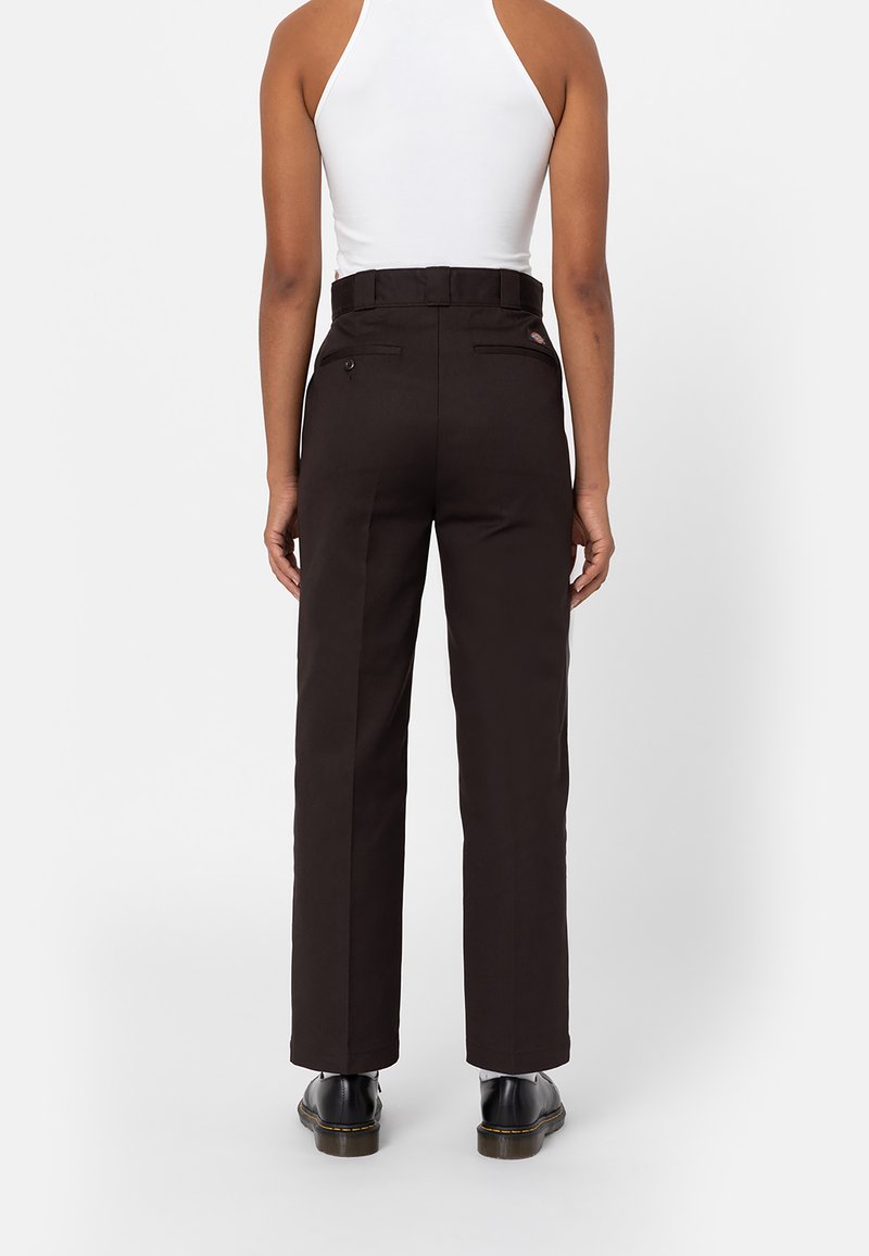Persona vestida con pantalones rectos negros, una blusa sin mangas blanca y zapatos negros, de pie con la espalda hacia la cámara frente a un fondo blanco.