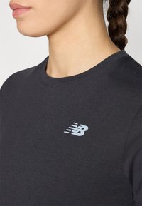 T-shirt noir à manches courtes en coton. Présente un logo New Balance bleu clair sur la poitrine, avec un col rond et une texture lisse.