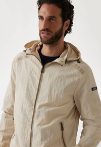 Light-colored, textured jacket with a zippered front, drawstring hood, and two side pockets. Features a brand patch on the sleeve.