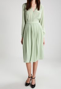 Hellgrünes Kleid mit langen Ärmeln, Knopfleiste, gesmoktem Rock und taillierter Passform. Getragen mit schwarzen High Heels mit Knöchelriemen.